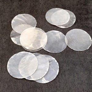 May include: A collection of translucent, round, flat, and irregularly shaped objects, possibly coasters or decorative elements. The objects are a pale, pearlescent color and are arranged on a dark, textured surface.