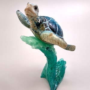May include: A sea turtle figurine with a dark blue and white patterned shell and light tan skin, positioned as if swimming above a translucent, bright green wave-shaped base that curves upward to support the turtle's body in mid-air.