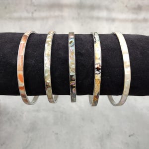 May include: Five silver bangles with various inlaid designs. The bangles feature a range of colors, including orange, white, and iridescent abalone shell. The bangles are displayed on a black surface.