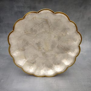 May include: A decorative, scalloped-edge serving tray. The tray features a pearlescent, white surface with a gold-colored trim. The tray's design is reminiscent of a flower, adding an elegant touch to any setting.