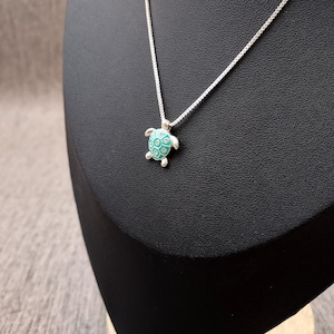 May include: A silver-toned chain necklace featuring a small turtle pendant. The turtle shell is accented with a bright turquoise enamel inlay and small, clear embedded stones arranged in a geometric pattern across the surface of the shell.