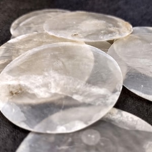 May include: A collection of translucent, iridescent, oval-shaped mother-of-pearl shells. The shells have a pearly white color with subtle variations in tone and texture. They are arranged in a slightly overlapping pattern, creating a sense of depth and layering.