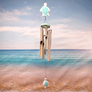 May include: A hanging wind chime with a sea turtle design. The wind chime features a white turtle at the top and bottom, with a turquoise design on the shell. The chimes are made of bamboo and hang in front of a beach scene.