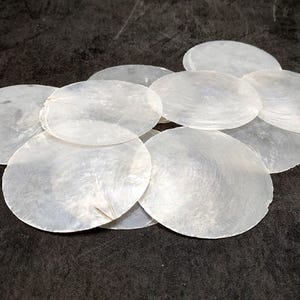 May include: A group of white, iridescent, round, mother of pearl shell pieces. The shells are arranged in a pile, with the largest shell at the bottom and the smaller shells stacked on top.