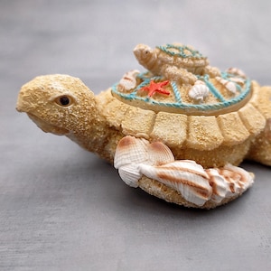 May include: A decorative turtle figurine in shades of beige and brown, adorned with seashells and a small turtle on its back. The turtle's shell features a blue rope design and a red starfish accent. A charming coastal-themed decorative item.