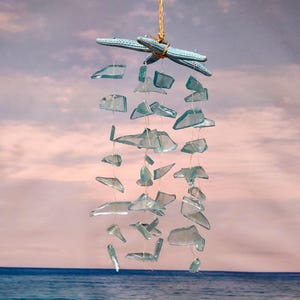 May include: A hanging wind chime featuring light blue sea glass pieces suspended from a natural rope. Two light blue starfish are tied to the top. The background shows a sky and ocean scene.