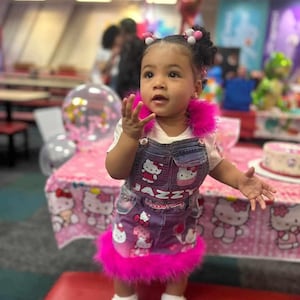 Puede incluir: Una niña pequeña con un vestido de peto vaquero con estampado de Hello Kitty rosa. El vestido tiene un ribete de plumas rosas y la niña lleva zapatillas blancas. Está de pie frente a una mesa con un mantel rosa de Hello Kitty.