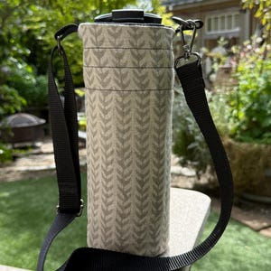 May include: A beige water bottle carrier with a black strap. The carrier has a repeating leaf pattern in a lighter beige color. The black strap is adjustable and has a metal clasp. The carrier is sitting on a wooden surface.