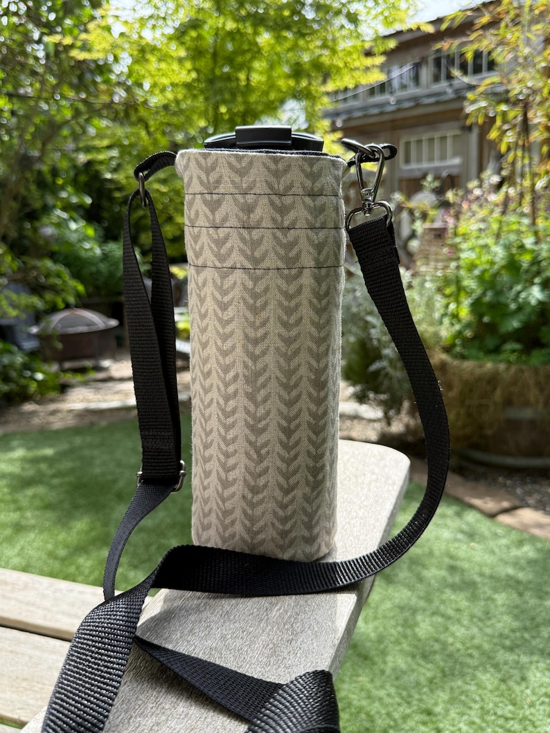 May include: A beige water bottle carrier with a black strap. The carrier has a repeating leaf pattern in a lighter beige color. The black strap is adjustable and has a metal clasp. The carrier is sitting on a wooden surface.