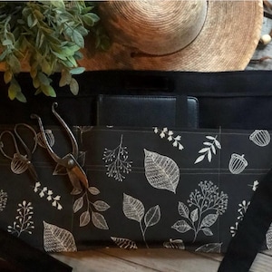 May include: A black waist apron featuring a white botanical print of leaves and acorns. The apron includes a solid black waistband with long ties and a large front pocket containing a black notebook and a pair of metal garden shears.