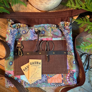 May include: A fabric bag featuring a colorful mandala pattern with a brown canvas front pocket. The pocket includes a leather strap with a brass clip, a pair of metal scissors, and two tan paper seed packets labeled GAURA WHIRLING BUTTERFLIES.