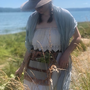 May include: A woman wearing a white dress with a lace trim, a light blue scarf, and a brown wide-brimmed hat. She is holding a bunch of green plants in her hand. She is standing in a field of tall grass.