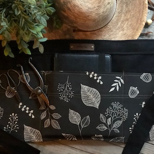 May include: Black gardening apron with multiple pockets, a white leaf pattern, and a black leather label. The apron is shown with gardening shears and a black notebook.