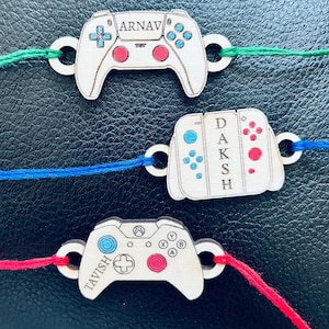 May include: Three wooden game controller-shaped ornaments with personalized names. Each controller has a different design and color scheme, with red, blue, and green accents. The names "ARNAV", "DAKSH", and "TAVISH" are engraved on each controller.