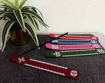 May include: A collection of colorful incense holders in red, blue, pink, and green, each adorned with white geometric patterns. The holders are rectangular and made of wood. An incense stick is lit and placed in the red holder.