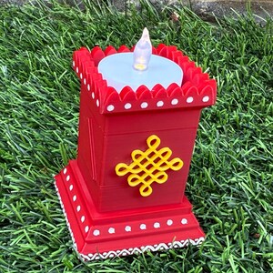 Pode incluir: Uma lanterna decorativa vermelha com uma vela LED branca. A lanterna tem uma forma quadrada com um design de nó amarelo e detalhes brancos. A lanterna está sobre a relva verde.