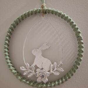 May include: An Easter wreath with a macrame ring in a pale green colour. A white metal egg shape holds a bunny and floral details. The wreath is designed for hanging, a charming seasonal decoration.
