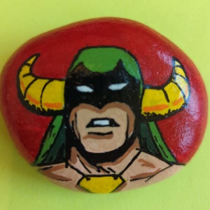 May include: Hand-painted rock art featuring a superhero with a green and yellow costume, black mask, and horns. The background is red.