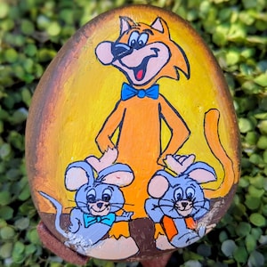 May include: Hand-painted, oval-shaped decorative stone featuring a cartoon cat in orange with a blue bow tie, and two gray mice. The background is yellow and orange, with a brown border.