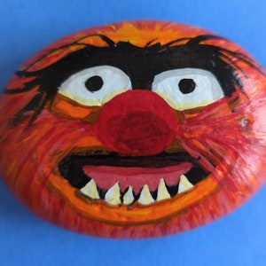 May include: Hand-painted rock with a red, orange, and black depiction of a furry monster with a big smile and a red nose.
