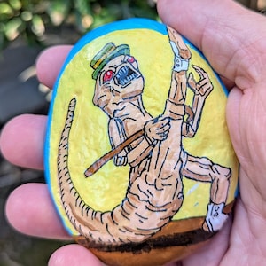 May include: Hand-painted rock art featuring a creature with red eyes, a green hat, and a long tail. The creature is painted on a yellow background with a blue border. The rock is held in a hand.