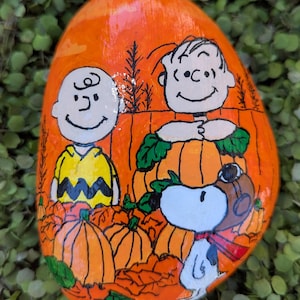 May include: A smooth, oval-shaped rock painted with a vibrant orange background featuring a hand-painted scene of Charlie Brown, Linus, and Snoopy in a pumpkin patch. The characters are depicted in black outlines with colorful details.