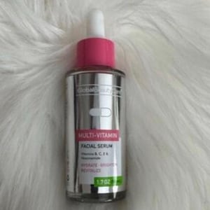 Może przedstawiać: Srebrna butelka serum do twarzy Global Beauty Multi-Vitamin z różową nakrętką i białą pipetą. Etykieta zawiera tekst "Multi-Vitamin Facial Serum" i "1.7 oz". Butelka znajduje się na białej, puszystej powierzchni.