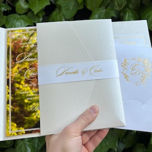 K&ouml;nnte beinhalten: Elfenbein-Hochzeitseinladungs-Set mit goldfarbenen Akzenten. Die Einladung zeigt ein florales Design und die Namen "Daniella & Carlos". Das Set enth&auml;lt eine Einladung, ein Bauchband und einen Umschlag.
