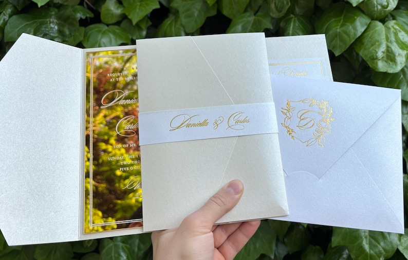 K&ouml;nnte beinhalten: Elfenbein-Hochzeitseinladungs-Set mit goldfarbenen Akzenten. Die Einladung zeigt ein florales Design und die Namen "Daniella & Carlos". Das Set enth&auml;lt eine Einladung, ein Bauchband und einen Umschlag.