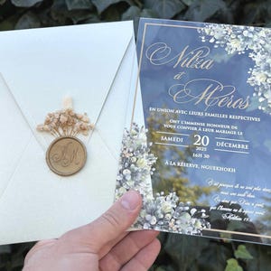 Könnte beinhalten: Eine transparente Acryl-Hochzeitseinladung mit goldener Schrift und Blumenmotiven sowie ein cremefarbener Umschlag mit einem goldenen Wachssiegel mit den Initialen MN, verziert mit einem kleinen Zweig getrockneter Schleierkrautblüten.