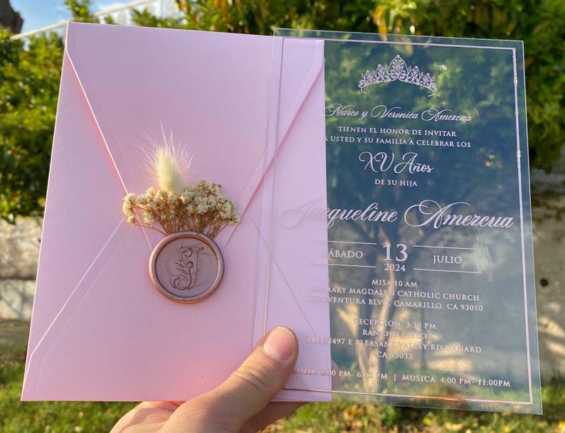 Pode incluir: Um convite transparente de acr&iacute;lico com letras cor-de-rosa para uma Quincea&ntilde;era. O convite inclui o texto "Marco y Veronica Amezcua tienen el honor de invitar a usted y su familia a celebrar los XV A&ntilde;os de su hija Jacqueline Amezcua" com a data "Sabado 13 Julio 2024".