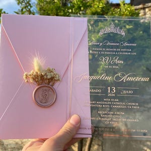 Pode incluir: Um convite de Quincea&ntilde;era rosa e dourado com um painel frontal de acr&iacute;lico transparente. O convite apresenta uma coroa dourada, o texto "Narco y Veronica Amezcua" e o texto "XV Aries". O convite tamb&eacute;m inclui a data "Sabado 13 Julio 2024", a hora "Misa 10 AM" e a localiza&ccedil;&atilde;o "St. Mary Magdalen Catholic Church".