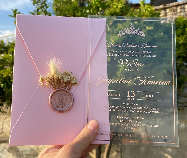 Pode incluir: Um convite de Quincea&ntilde;era rosa e dourado com um painel frontal de acr&iacute;lico transparente. O convite apresenta uma coroa dourada, o texto "Narco y Veronica Amezcua" e o texto "XV Aries". O convite tamb&eacute;m inclui a data "Sabado 13 Julio 2024", a hora "Misa 10 AM" e a localiza&ccedil;&atilde;o "St. Mary Magdalen Catholic Church".