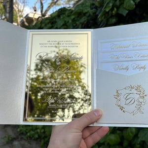 K&ouml;nnte beinhalten: Eine cremefarbene Hochzeitseinladung mit goldenen Akzenten. Die Einladung hat eine verspiegelte Mittelplatte mit eleganter Schrift und eine Tasche mit kleineren Karten. Das Design umfasst florale Elemente und den Text "Kindly Reply".