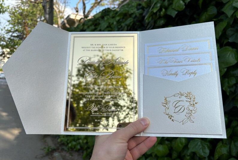K&ouml;nnte beinhalten: Eine cremefarbene Hochzeitseinladung mit goldenen Akzenten. Die Einladung hat eine verspiegelte Mittelplatte mit eleganter Schrift und eine Tasche mit kleineren Karten. Das Design umfasst florale Elemente und den Text "Kindly Reply".