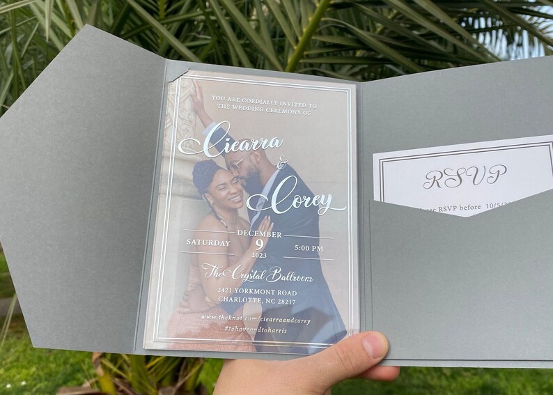 May include: A wedding invitation with a clear plastic cover featuring a photo of a couple. The invitation is for Cicarra and Corey's wedding on December 9, 2023, at 5:00 PM at The Crystal Ballroom. The invitation includes the address and website for the event.