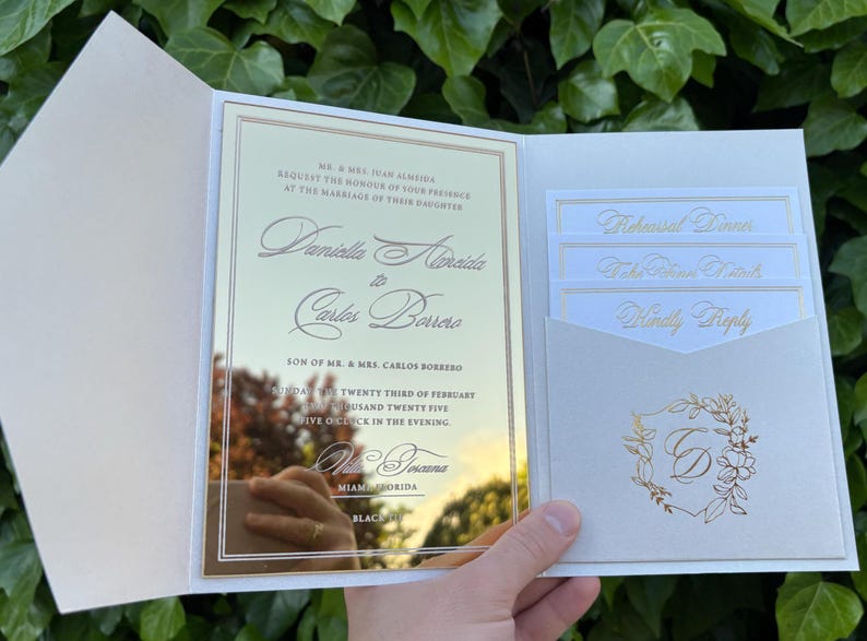 K&ouml;nnte beinhalten: Ge&ouml;ffnete Hochzeitseinladungs-Suite mit goldener Spiegelkarte und cremefarbenen Eins&auml;tzen. Die Hauptkarte zeigt elegante Schrift mit Namen und Veranstaltungsdetails. Zus&auml;tzliche Karten enthalten "Rehearsal Dinner", "RSVP" und ein Blumenschild.