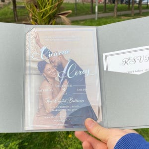 May include: A gray wedding invitation with a clear plastic insert featuring a photo of a couple. The text on the invitation reads "Cicaru & Corey" and includes the date and time of the wedding. The RSVP card is in a pocket on the right side of the invitation.