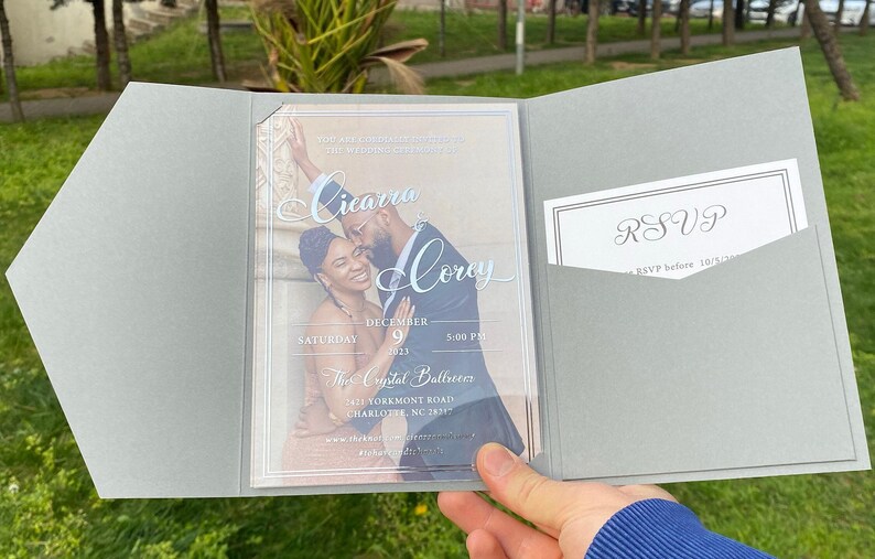 May include: A gray wedding invitation with a clear plastic insert featuring a photo of a couple. The text on the invitation reads "Cicaru & Corey" and includes the date and time of the wedding. The RSVP card is in a pocket on the right side of the invitation.