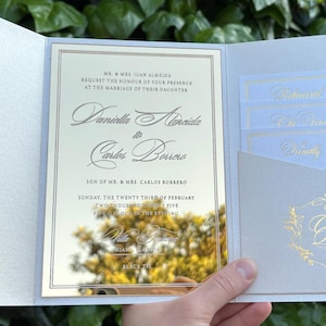 K&ouml;nnte beinhalten: Elegante Hochzeitseinladung mit hellblauem und goldenem Design. Die Einladung zeigt die Namen des Paares, das Datum und die Uhrzeit der Zeremonie. Enth&auml;lt eine Tasche f&uuml;r RSVP-Karten und ein dekoratives Monogramm.
