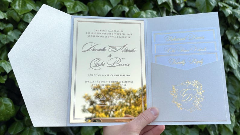 K&ouml;nnte beinhalten: Elegante Hochzeitseinladung mit hellblauem und goldenem Design. Die Einladung zeigt die Namen des Paares, das Datum und die Uhrzeit der Zeremonie. Enth&auml;lt eine Tasche f&uuml;r RSVP-Karten und ein dekoratives Monogramm.
