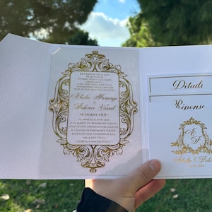 Pode incluir: Convite de casamento branco com detalhes dourados ornamentados. O convite inclui texto em francês, com os nomes Elodie Mesnage e Fabrice Vassal. Há um bolso para detalhes e cartões de resposta.