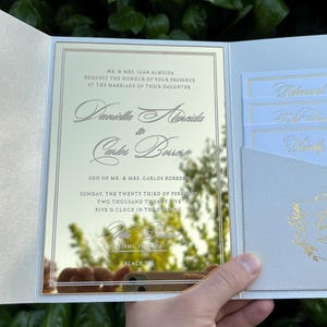 K&ouml;nnte beinhalten: Eine cremefarbene Hochzeitseinladung mit goldfarbenen Buchstaben und einer reflektierenden Mittelplatte. Die Einladung zeigt die Namen des Paares und die Veranstaltungsdetails. Zus&auml;tzliche Eins&auml;tze sind sichtbar.