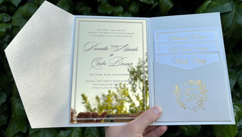 K&ouml;nnte beinhalten: Eine cremefarbene Hochzeitseinladung mit goldfarbenen Buchstaben und einer reflektierenden Mittelplatte. Die Einladung zeigt die Namen des Paares und die Veranstaltungsdetails. Zus&auml;tzliche Eins&auml;tze sind sichtbar.