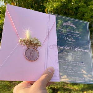 Pode incluir: Um convite de Quincea&ntilde;era em acr&iacute;lico rosa e transparente. O convite apresenta uma coroa, o texto "Mares y Veronica Amezcua", "XV A&ntilde;os", "Jacqueline Amezcua" e a data "Sabado 13 Julio". O convite tamb&eacute;m inclui o local e a hora do evento.