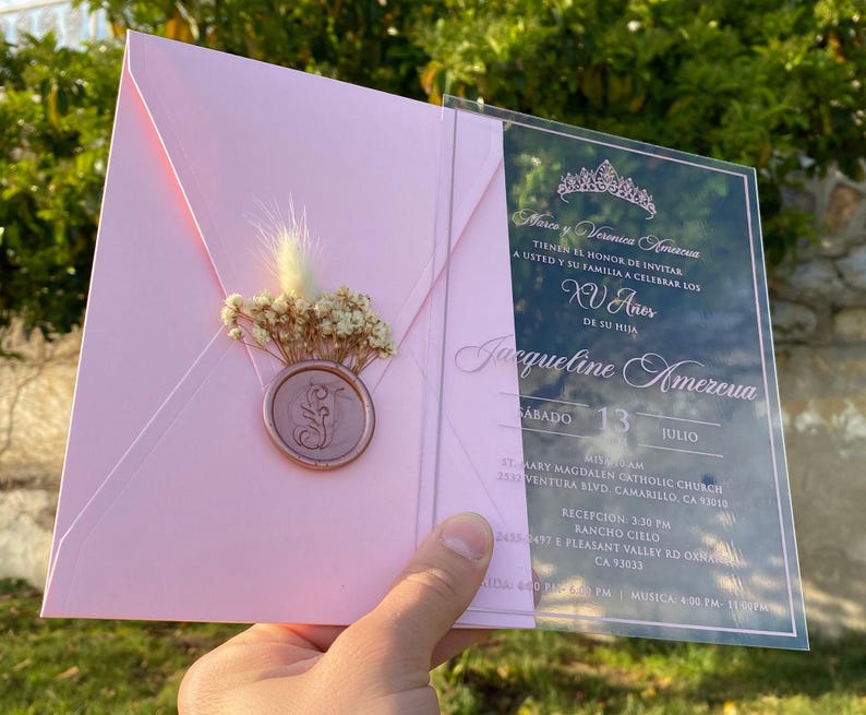 Pode incluir: Um convite de Quincea&ntilde;era em acr&iacute;lico rosa e transparente. O convite apresenta uma coroa, o texto "Mares y Veronica Amezcua", "XV A&ntilde;os", "Jacqueline Amezcua" e a data "Sabado 13 Julio". O convite tamb&eacute;m inclui o local e a hora do evento.