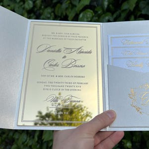 K&ouml;nnte beinhalten: Elegante Hochzeitseinladungssuite mit Perlmutt-Finish. Die Einladung hat goldfarbenen Text auf einer verspiegelten Oberfl&auml;che, mit zus&auml;tzlichen Karten in einer Tasche. Die Suite enth&auml;lt die Namen des Paares und Hochzeitsdetails.