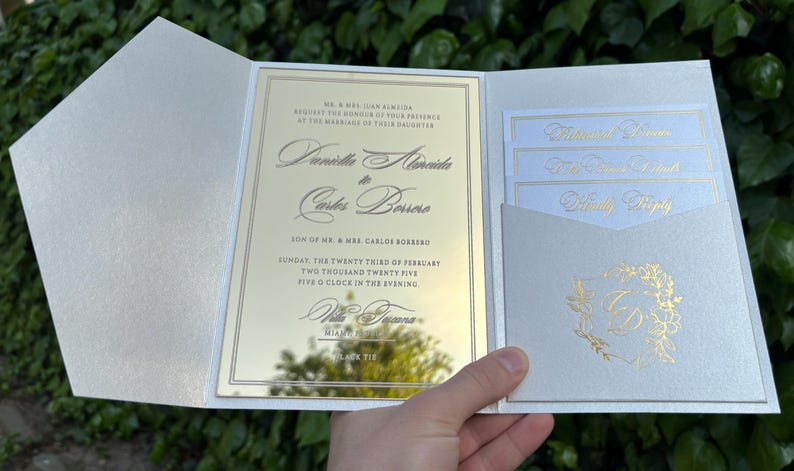 K&ouml;nnte beinhalten: Elegante Hochzeitseinladungssuite mit Perlmutt-Finish. Die Einladung hat goldfarbenen Text auf einer verspiegelten Oberfl&auml;che, mit zus&auml;tzlichen Karten in einer Tasche. Die Suite enth&auml;lt die Namen des Paares und Hochzeitsdetails.