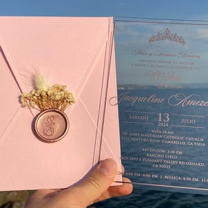 Pode incluir: Um convite de quincea&ntilde;era em acr&iacute;lico rosa e transparente. O convite apresenta uma coroa, o nome Jacqueline Amezcua e a data de s&aacute;bado, 13 de julho de 2024. O convite tamb&eacute;m inclui detalhes sobre a missa e a recep&ccedil;&atilde;o.