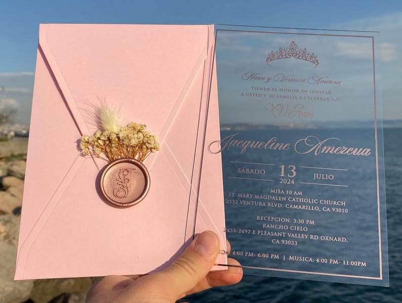 Pode incluir: Um convite de quincea&ntilde;era em acr&iacute;lico rosa e transparente. O convite apresenta uma coroa, o nome Jacqueline Amezcua e a data de s&aacute;bado, 13 de julho de 2024. O convite tamb&eacute;m inclui detalhes sobre a missa e a recep&ccedil;&atilde;o.